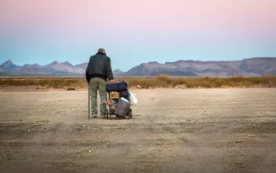 Older unhoused man standing next to cart with belongings and a cardboard sign "West" in a desert landscape.