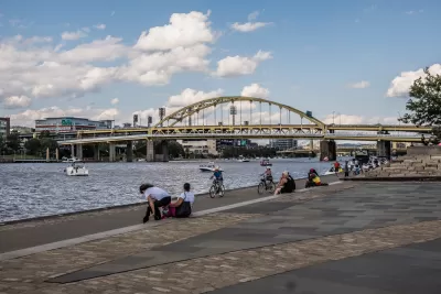People on bikes and sitting on ledge on Pittsburgh riverfront with arched bridge in background