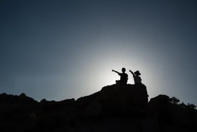 Two children sitting on rock sillhouetted against sky.