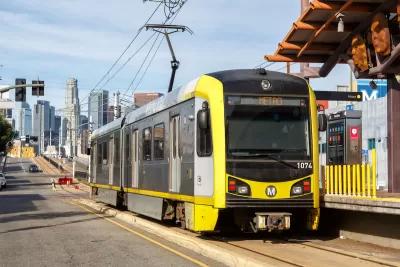 Yellow and black light rail train at street-level station with downtown Los Angeles skyline in background.