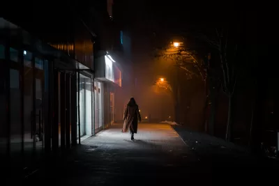 Silhouette of woman in long coat walking on darkened city street alone at night, passing brightly lit sotrefront.