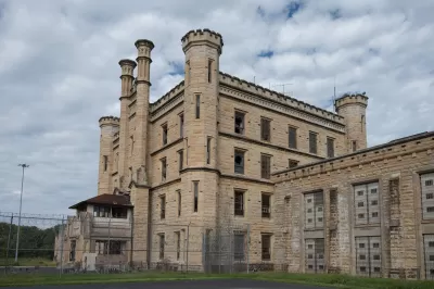 Deralict stone prison building with turrets and a chain link fence in foreground, under a gray, cloudy sky.