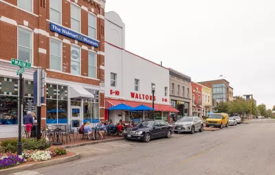 Main Street in Bentonville, Arkansas with three-story brick buildings, people sitting at tables on sidewalk, and sign on building for "The Walmart Museum."