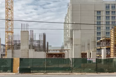 Multistory concrete apartment building under construction behind green construction fence.