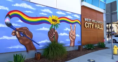 Front entrance of West Hollywood City Hall with name on side of building and mural of hands painting a rainbow across a blue sky.