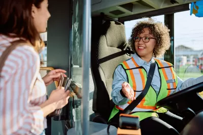 Female bus driver wearing safety vest sits in driver's seat, talking with woman passenger boarding bus
