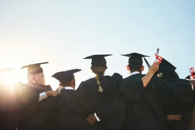 View from back of group of college students in caps and gowns at graduation with bright sunlight in front of them.