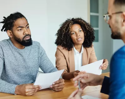 A Black couple holding contracts sits across the desk from a white mortgage lender.