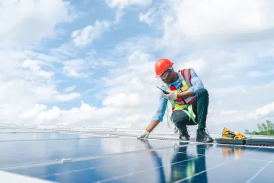 Worker wearing orange hard hat and vest over light blue long-sleeved shirt and jeans inspects rooftop solar panel