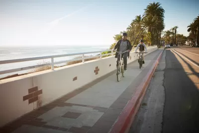 Father and son riding bikes on a sidewalk next to a road withough bike lane on California coast with palm trees in background at golden hour.