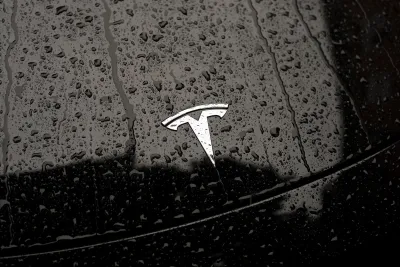 Close-up on silver Tesla logo on black car hood dotted with raindrops.