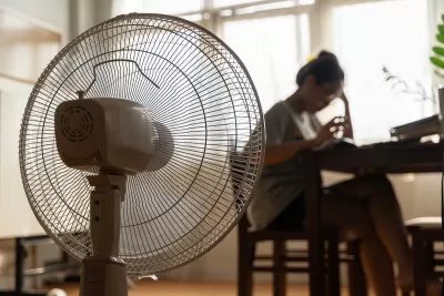 Close-up of floor fan operating with blurred woman sitting at table in background.