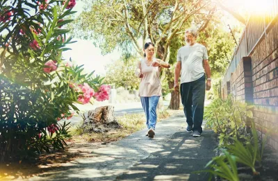 Older man and woman walking down sidewalk landscaped with bougainvillas next to a brick wall on a sunny day.