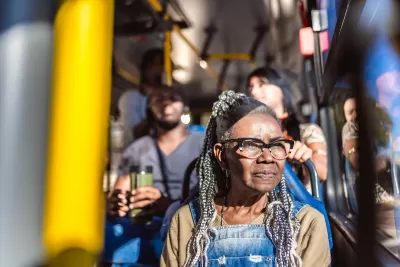 Older Black woman with glasses and braids sitting on city bus, looking out window.