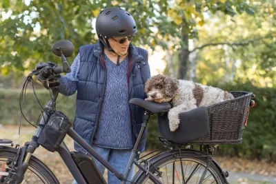 ELderly woman wearing black helmet and blue puffy vest stands next to e-bike with white and brown dog in rear basket.
