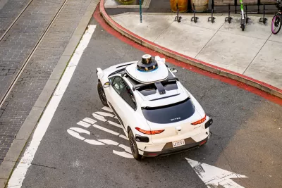 Overhead view of white Waymo self-driving car in San Francisco, California with shared bikes and scooters visible on adjacent sidewalk.