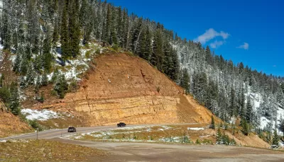 A road winds along the edge of a mountain, with an inclined tan rockface above it and a snowy treeline near the top of a photo.