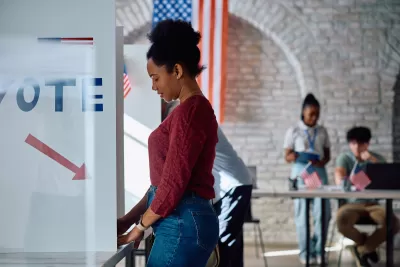 A side view of a young Black woman in a red sweater and jeans standing at a ballot box in a gymnasium with white walls and an American flag in the background.