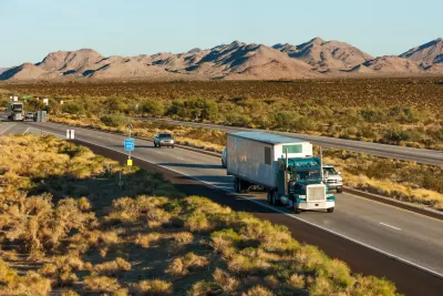 Double-lane interestate in the desert with two semi-trucks and five cars driving toward the camera, low mountains in the background.