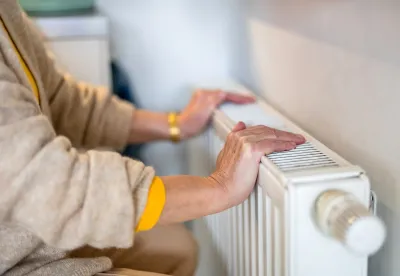 Close-up of older woman's hands resting on white modern heating radiator mounted on wall indoors.