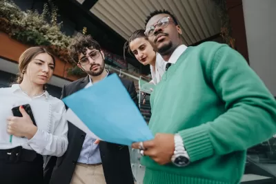Group of four young professional people standing in front of urban building looking at documents.