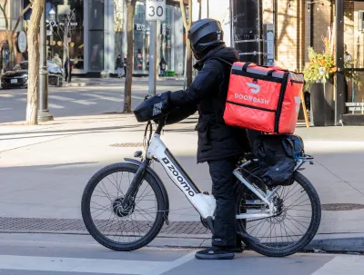 Person in black clothing and helmet with red DoorDash box on back of electric bike on urban street.