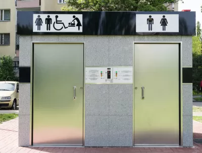 Outdoor public restrooms with two silver doors and icons for men, women, wheelchair accessibility, and baby changing above doors.