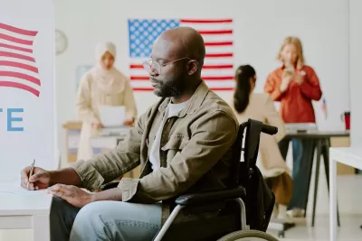 African American man in a wheelchair filling out paper ballot at a polling station with American flags in background.