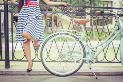 Close-up on woman in white and blue striped knee-length dress standing next to mint green cruiser bike resting against low wrought iron fence in front of green lawn.