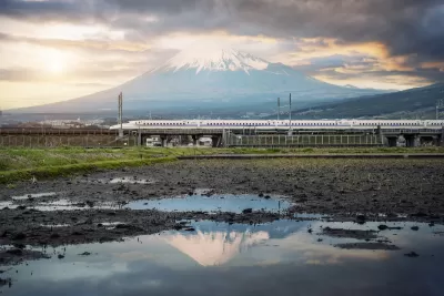 White Shinkansen high-speed rail train passing through green fields with water and Mount Fuji in background in Japan.