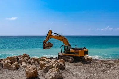 Yellow excavator on rocky beach with bright blue water in background.