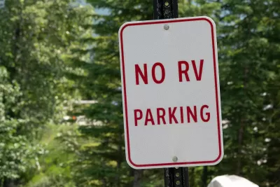 Close-up of white sign with red text 'No RV parking' against green foliage.