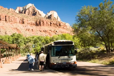 White shuttle bus picks up passengers in Zion National Park, red cliffs and mountains in background.