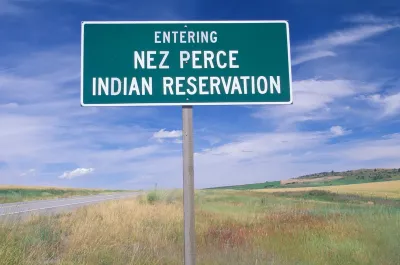 Green roadside sign with white text reading "Entering Nez Perce Indian Reservation" against grassy field and blue sky.