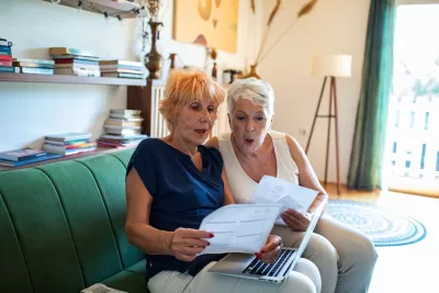 Two older women sitting on green sofa looking at paperwork or bills.