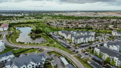 Aerial view of suburban mixed-use development outside Austin, Texas with lake in foreground and complex of apartment buildings.
