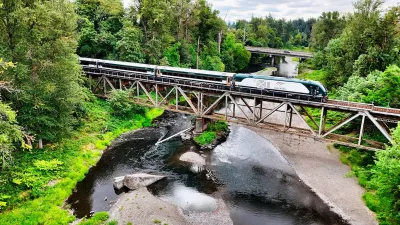 An Airo train passes over a mostly-dried river in between two large forests.
