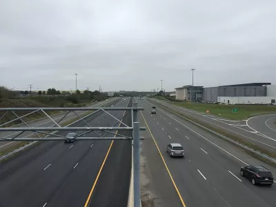 A wide highway with a few cars on a gloomy day. Low-rise commercial buildings and more car infrastructure is visible to the side of the road.