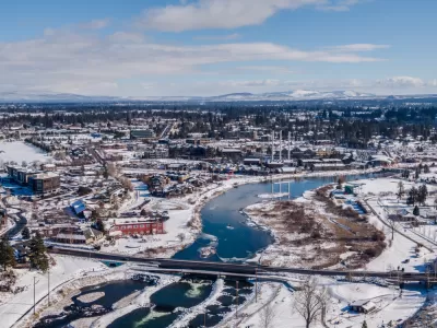 An aerial image of Bend, Oregon, covered in snow in the winter. The Deschutes River cuts through the middle of town, below an old mill.