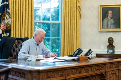 President Jose Biden appears in a collared, button down shirt while signing papers while seated at the desk in the Oval Office of the White House