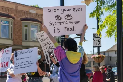 A Black LIves Matter Protest, with a protestor holding up a sign that reads "Open Your Eyes: Systemic Racism Is Real"