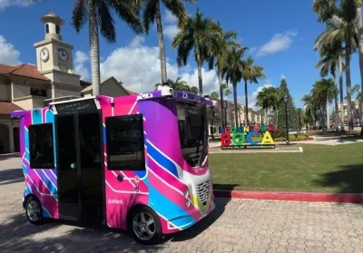 Colorful pink and purple small autonomous transit shuttle in park in Boca Raton, Florida with tall palm trees lining lawn in background.
