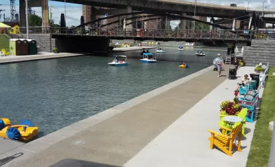 A canal under a bridge in Buffalo is lined by chairs and tables, people walking about, looking at peddle boats on the water.