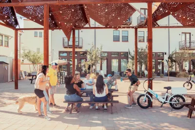 Group of young people sitting at outdoor shaded table laughing and talking with e-bikes and dog next to them.