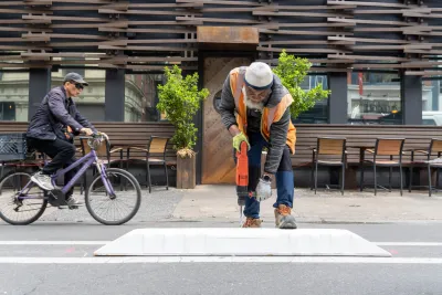 Worker in orange vest installing plastic bike lane separator as man rides past on bicycle in background.