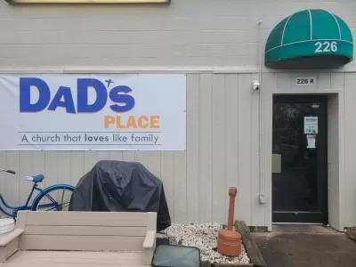 White sign on side of tan building with metal siding and green overhang, reading "Dad's Place, a church that loves like family."