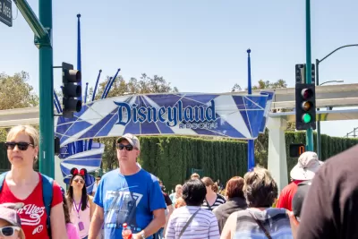 A crowd of people cross the street with a sign for the entrance of Disneyland in the background