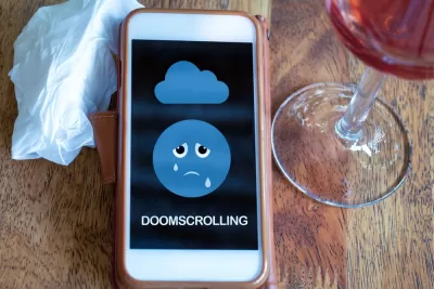 A mobile phone with a crying emoji face and the word "doomscrolling" is on a table between a tissue and a glass of wine.
