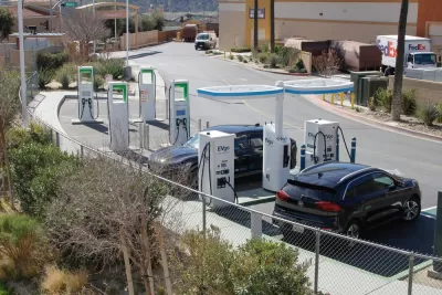 A pair of electric cars plugged into charge at facilities in the corner of a shopping center parking lot.