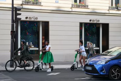 A woman on an electric bike and two women on electric scooters wait at an intersection for the light to change.